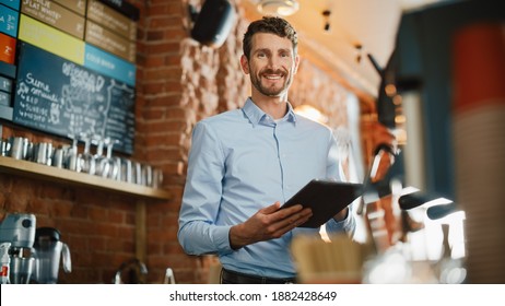 Handsome Caucasian Coffee Shop Owner is Working on Tablet Computer and Checking Inventory in a Cozy Loft-Style Cafe. Successful Restaurant Manager Standing Happy Behind Counter and Smiles on Camera.