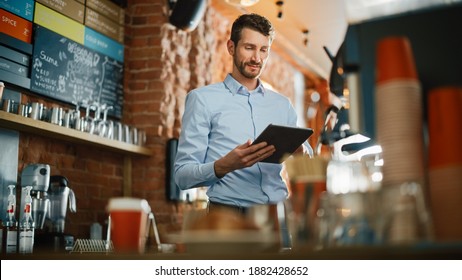 Handsome Caucasian Coffee Shop Owner Is Working On Tablet Computer And Checking Inventory In A Cozy Loft-Style Cafe. Successful Restaurant Manager Standing Happy Behind Counter.