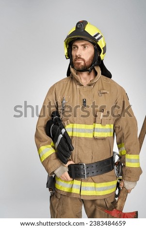 Handsome caucasian firefighter in helmet looking away, while carrying fire ax. Portrait of bearded man in fire fighting uniform with hand on belt posing, isolated on gray. Concept of work, profession.