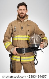 Handsome caucasian firefighter smiling to camera indoors. Portrait of happy, strong male fireman in protective uniform holding helmet, on white studio background. Workplace, safety concept.