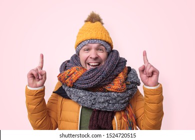 Handsome Caucasian Funny Man In Several Hats And Scarfs Pointing With Index Finger Up On Pink Background. Winter Fashion.