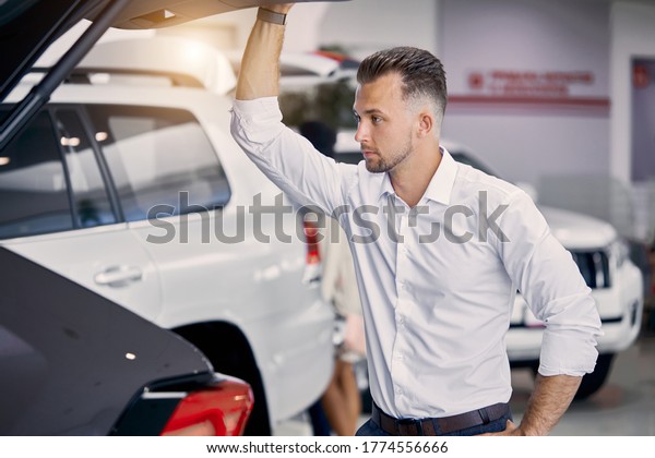 handsome\
caucasian guy examine the trunk of car in dealership , he looks\
inside of car from back and think buy it or\
not