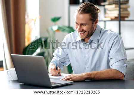 Handsome caucasian guy freelancer uses laptop. Smiling attractive modern man in stylish casual wear sits at the desk in a creative or home office, browsing internet, replies to email, distant learners
