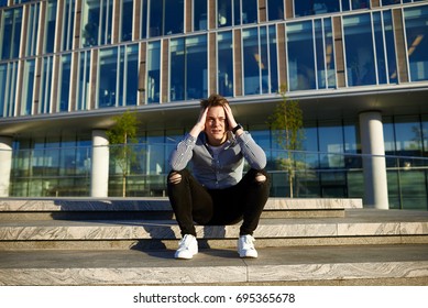 Handsome Caucasian Guy Wearing White Sneakers, Ripped Jeans And Shirt Sitting On Concrete Stairs In Urban Setting, Holding Head, Feeling Unhappy And Stressed Out After Big Fight With His Girlfriend