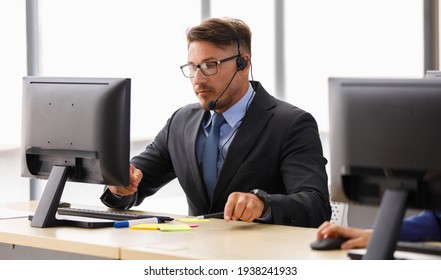 Handsome Caucasian Happy Male Operator Wearing Black Suit White Shirt And Blue Necktie Sit Smiling To Camera And Working Receiving Call By Headphone Microphone Behind Big Monitor In Office Room.