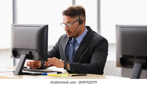 Handsome Caucasian Happy Male Operator Wearing Black Suit White Shirt And Blue Necktie Sit Smiling To Camera And Working Receiving Call By Headphone Microphone Behind Big Monitor In Office Room.