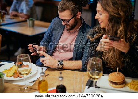 Handsome caucasian hipster man taking photo of his food while sitting in restaurant. Next to him is his girlfriend holding smart phone and looking at him.