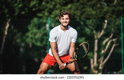 A handsome Caucasian male tennis player smiling, carrying his racket and ready to play on an outdoor court, with trees in the background. Concept of joy of playing sports and recreation outdoors. - Powered by Shutterstock