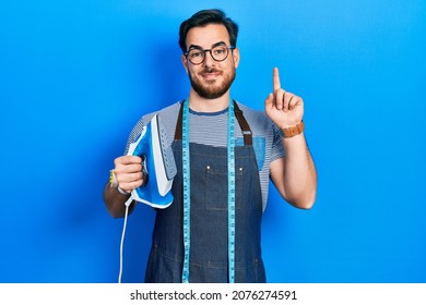 Handsome caucasian man with beard dressmaker designer wearing atelier apron holding iron surprised with an idea or question pointing finger with happy face, number one 