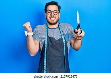 Handsome Caucasian Man With Beard Dressmaker Designer Wearing Atelier Apron Holding Scissors Screaming Proud, Celebrating Victory And Success Very Excited With Raised Arm 