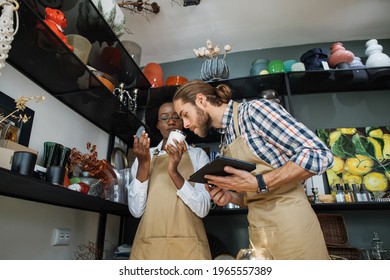 Handsome Caucasian Man With Digital Tablet In Hands Smelling Aroma Candle That Holding African Saleswoman. Two Multiracial Partners In Apron Doing Inventory At Decor Store.