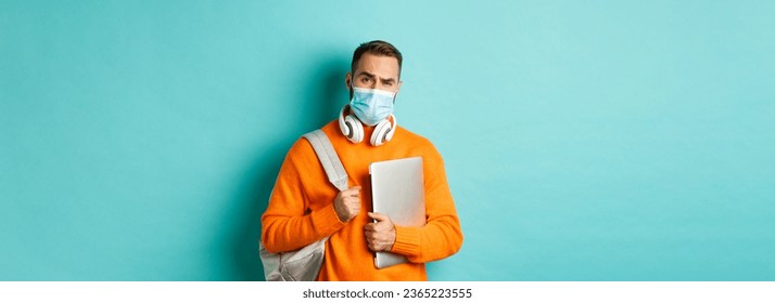 Handsome caucasian man with headphones and backpack, holding laptop and wearing medical mask, looking doubtful and skeptical, standing over light blue background. - Shutterstock ID 2365223555