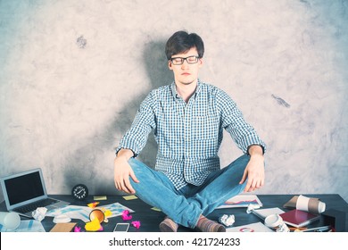 Handsome Caucasian Man Meditating On Messy Desktop With Blank Laptop Screen On Concrete Wall Background. Mock Up