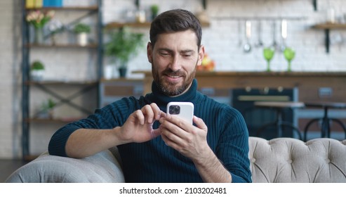 Handsome caucasian man sitting on sofa at cosy home and using smartphone surfing internet social media, watching videos and smiling.  - Powered by Shutterstock