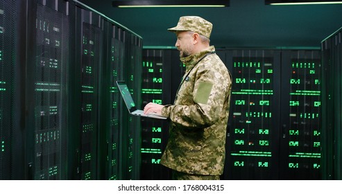 Handsome Caucasian Man From US Army Typing On Keyboard Of Laptop Computer While Checking Data At Servers Monitors In Dark Room. Male Technician Of Anti-terrorism Center.