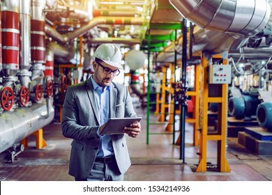 Handsome Caucasian Manager In Gray Suit And With Helmet On Head Using Tablet While Standing In Power Plant.