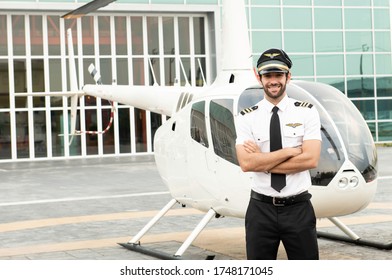 Handsome Caucasian Pilot In White Shirt Uniform And Hat Standing Folded Hand At Helicopter Platform Background.Handsome Young White Man Pilot With Private Motor Airplane Brackground. Ready To Flight.