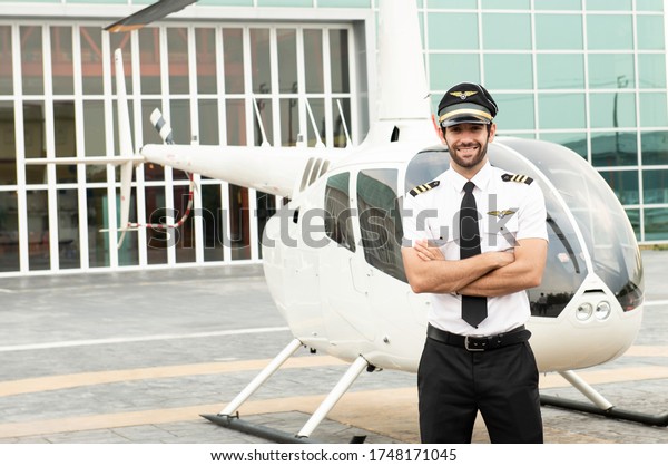 Handsome caucasian pilot in white shirt uniform\
and hat standing folded hand at helicopter platform\
background.Handsome young white man pilot with private motor\
airplane brackground. Ready to\
flight.