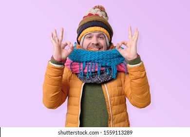 Handsome Caucasian Shocked Funny Man In Several Hats And Scarfs Showing Ok Gesture On Colored Background. Winter Fashion.