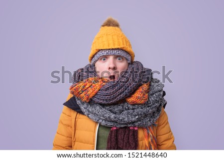 Handsome caucasian shocked funny man in several hats and scarfs on colored background. Winter fashion.