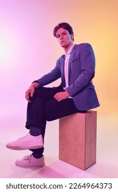 Handsome caucasian young man  business man in jacket   jeans sitting wooden box over light gradient yellow  purple background  Fashion  art  education  business concept  Looks thoughtful