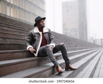 A handsome and charismatic black man in a black hat, sheepskin coat and black jeans sits on the steps. Stylish man.
