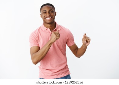 Handsome charming young african american masculine guy in pink polo shirt gesturing with hands as dancing, having fun smiling joyfully spending great time at friends party over white wall