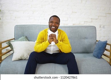 Handsome cheerful dark skinned man wearing jeans and yellow cardigan, sitting on comfortable sofa in living room, clasping hands and smiling broadly at camera, showing his white perfect teeth - Shutterstock ID 1120427861