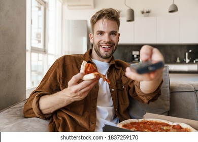 Handsome Cheerful Guy Eating Pizza While Watching Tv And Using Remote Control At Home