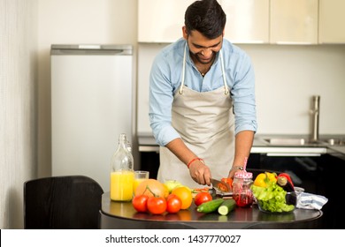 Handsome Cheerful Smiling Indian Man Cooking Summer Salad For Dinner, Cutting Tomato On Chopping Board, Preparing Dinner For Wife At Home In The Kitchen. Healthy Food, Cooking Concept.