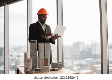 Handsome chief architect african man in business suit and protective helmet with laptop pc working with city buildings model as urban planner at office interior with panoramic city view.