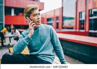 Handsome Chinese Hipster Guy Dressed In Casual Wear Calling To Service Via Smartphone Device Sitting At Urban Setting, Youthful Japanese Male Using Roaming Internet Connection For Cellular Phoning