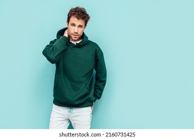 Handsome confident hipster  model.Sexy unshaven man dressed in summer stylish green hoodie and jeans clothes. Fashion male with curly hairstyle posing in studio. Isolated on blue - Shutterstock ID 2160731425