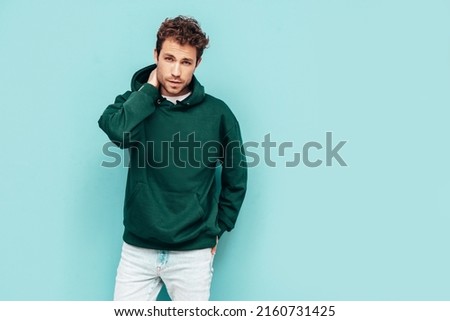 Handsome confident hipster  model.Sexy unshaven man dressed in summer stylish green hoodie and jeans clothes. Fashion male with curly hairstyle posing in studio. Isolated on blue