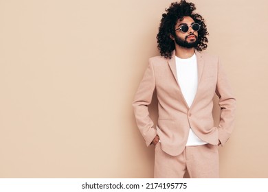 Handsome confident hipster  model.Unshaven Arabian man dressed in elegant beige suit . Fashion male with long curly hairstyle posing near beige wall in studio. Isolated. In sunglasses