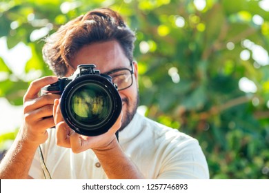 handsome and confident indian man photographer with a large professional camera taking pictures photo shooting on the beach.photo session on summer holiday on the background of green tropical trees