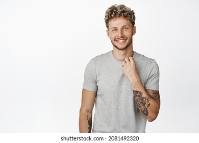 Handsome and confident macho man with perfect white smile, adjusting his t-shirt collar and smiling self-assured at camera, standing over white background