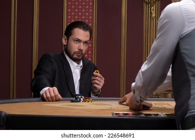 Handsome  confident man   in casino looking at  dealer  sitting at table with  chips and cards waist up