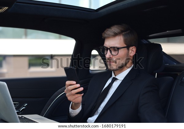 Handsome confident man in\
full suit looking at his smart phone while sitting in the car and\
using laptop.