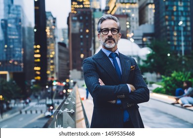 Handsome Confident Middle Aged Male In Glasses And Business Suit With Arms Crossed Standing Near Glass Fence Against Evening New York Street