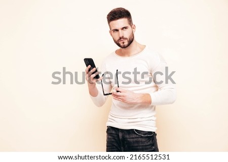 Handsome confident model.Sexy stylish man dressed in sweater and jeans. Fashion hipster male posing near white wall in studio. Holding smartphone. Looking at cellphone screen. Using apps