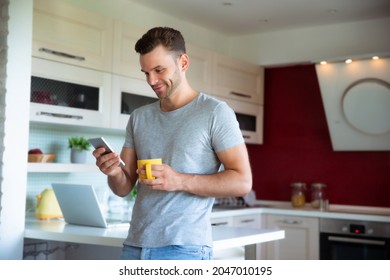 Handsome confident modern man in gray t-shirt stands with phone in hands and drinks coffee from the yellow cup on the kitchen at home - Powered by Shutterstock