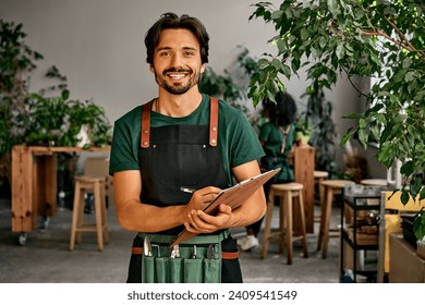 Handsome confident sincere smiling male gardener florist in green t-shirt black apron and bag of garden tools on belt holding wooden clipboard and pen looking at camera. Work in a plant shop. - Powered by Shutterstock