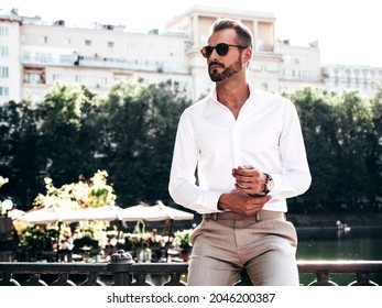 Handsome confident stylish hipster lambersexual model.Sexy modern man dressed in white shirt and trousers. Fashion male posing in the street near embankment in Europe park city at sunset.In sunglasses - Shutterstock ID 2046200387