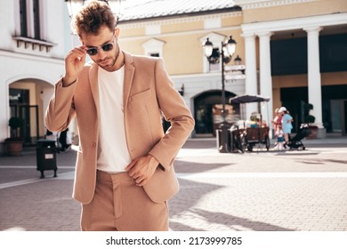 Handsome confident stylish hipster lambersexual model.Sexy modern man dressed in elegant beige suit. Fashion male posing in the street background in Europe city at sunset. In sunglasses