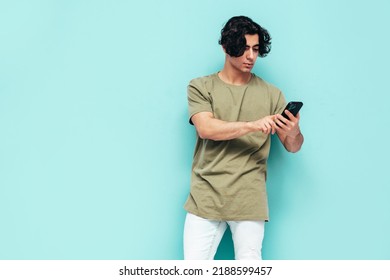 Handsome confident  stylish hipster lambersexual model.Man dressed in over size T-shirt and jeans. Fashion male posing near blue wall.Holding smartphone. Looking at cellphone screen. Using apps - Shutterstock ID 2188599457