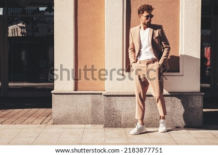 Handsome confident stylish hipster lambersexual model.Sexy modern man dressed in elegant beige suit. Fashion male posing in the street background in Europe city at sunset. In sunglasses. Full length