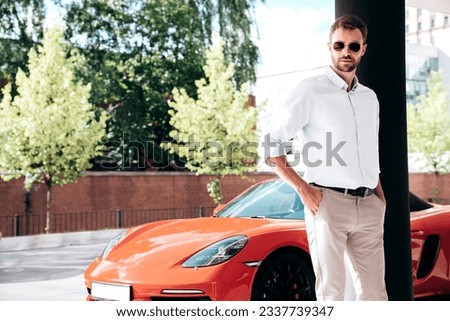 Handsome confident stylish hipster lambersexual model. Modern man dressed in white shirt and trousers. Fashion male posing in the street urban background in sunglasses. Outdoors. Near car, luxury life
