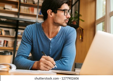 Handsome confident young man sitting at the library desk, working/studying, using laptop computer - Shutterstock ID 1277902606