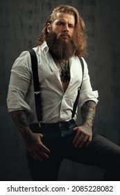 Handsome Courageous Middle-aged Man With Long Hair And A Thick Beard Posing In The Studio In Jeans And A Shirt With Suspenders